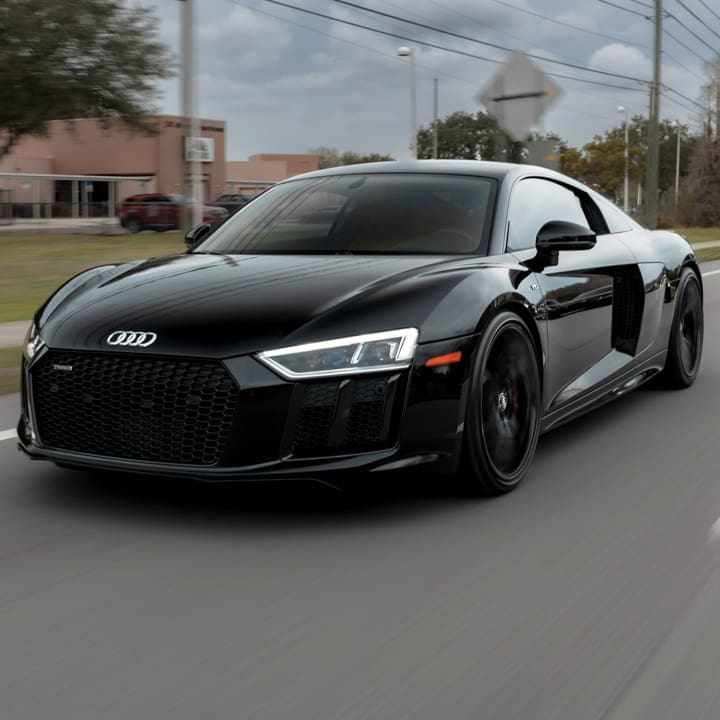 Black Audi R8 driving on a road, front view with visible grille and headlights.