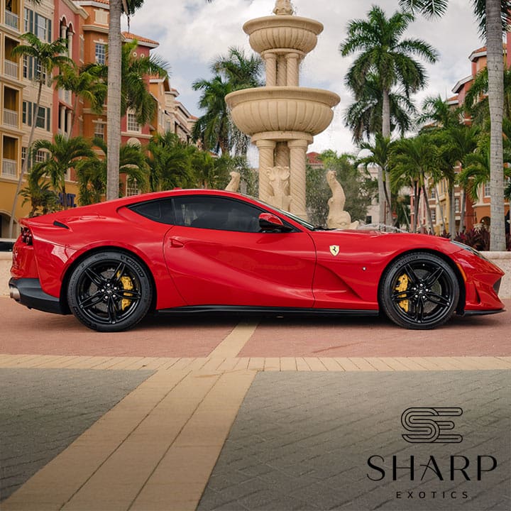 Red sports car parked in front of a large fountain, surrounded by palm trees and colorful buildings, with Sharp Exotics logo in corner.