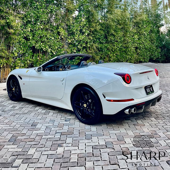 White convertible sports car parked on a brick driveway, surrounded by lush green foliage.