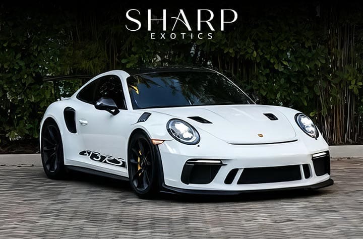 White sports car with black accents parked on a cobblestone surface, surrounded by greenery, featuring "Sharp Exotics" text above.