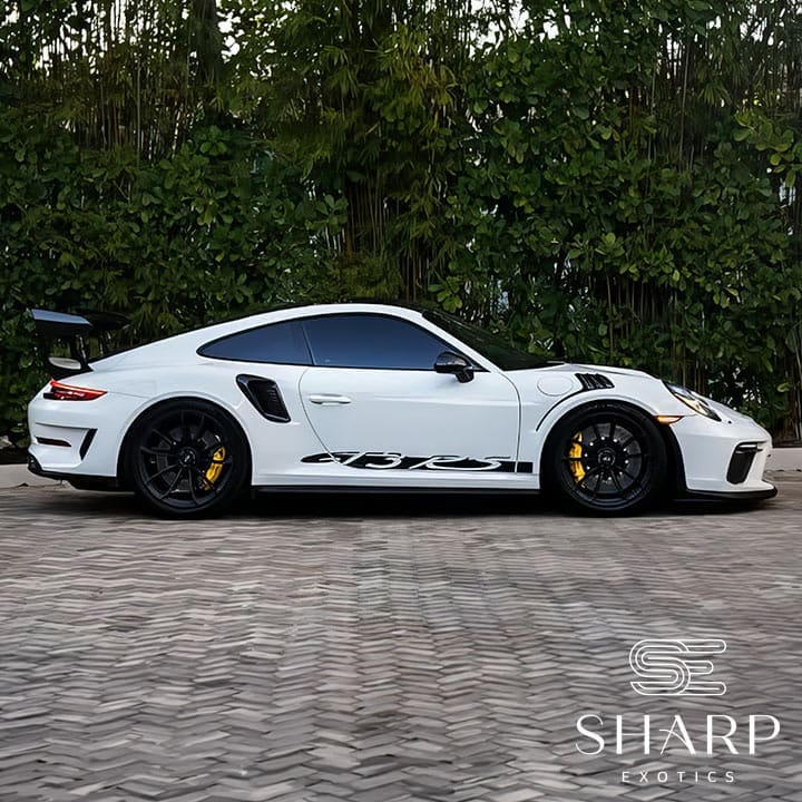White sports car with black accents and a rear spoiler, parked on a brick surface with greenery in the background.