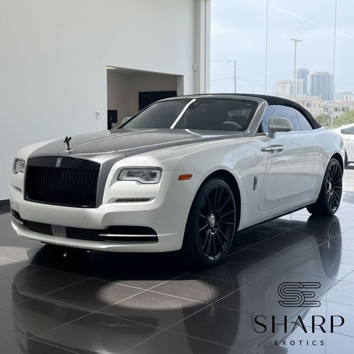 White luxury convertible car with black grille and wheels, parked indoors on a glossy floor. Logo reads "Sharp Exotics" in the corner.