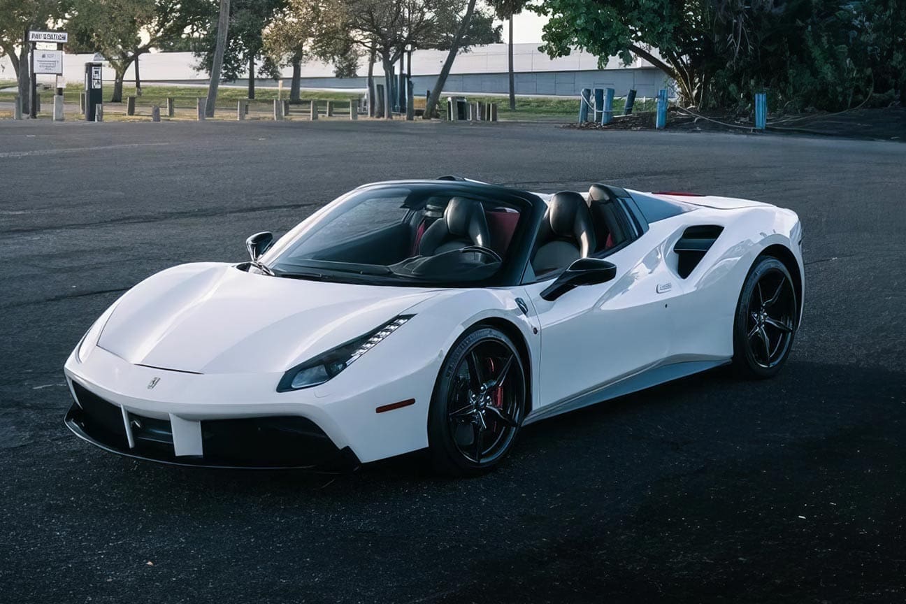 White convertible sports car parked in an empty lot with trees and a pay station in the background.