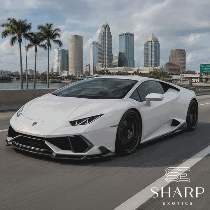 White sports car driving on a highway with a city skyline and palm trees in the background, logo "Sharp Exotics" in the corner.