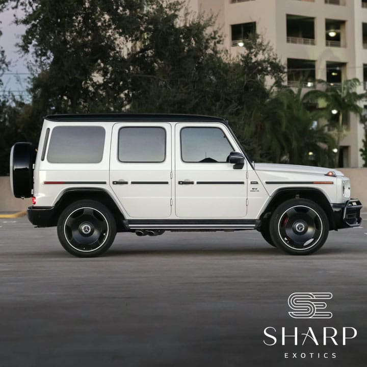 White SUV parked in an urban setting with trees and a building in the background, featuring the Sharp Exotics logo.
