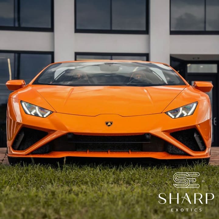 Orange sports car parked on grass, front view, with a building in the background and "Sharp Exotics" logo in the corner.