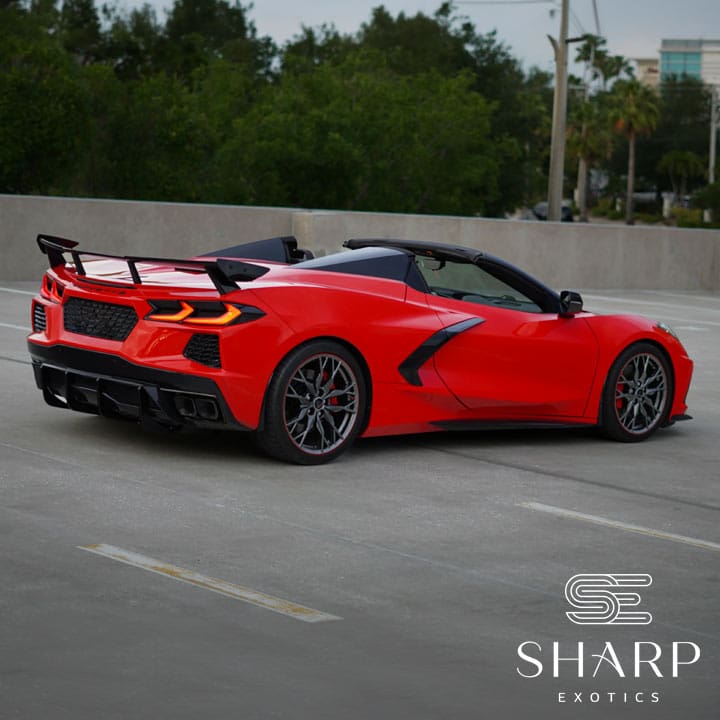 Red sports car with a rear spoiler parked on a rooftop, surrounded by trees and buildings, featuring the Sharp Exotics logo.
